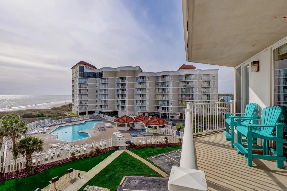 This exterior hero image showcases the condo’s private balcony with inviting chairs, overlooking a sparkling saltwater pool, lush landscaped gardens, and an expansive ocean view beyond. The tranquil scene hints at sunbathing and socializing by the pool, watching sunrises over the Atlantic, and relaxing evenings stargazing from the comfort of your balcony.