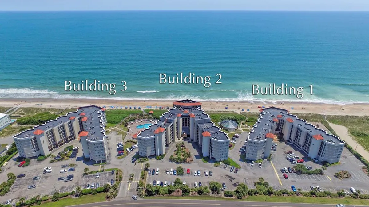 This image displays three residential buildings directly on the beachfront, with a swimming pool in the foreground. The scene includes the sandy beach, the shimmering Atlantic Ocean, and sunlit balconies facing the water, with inviting poolside loungers and a sense of community.