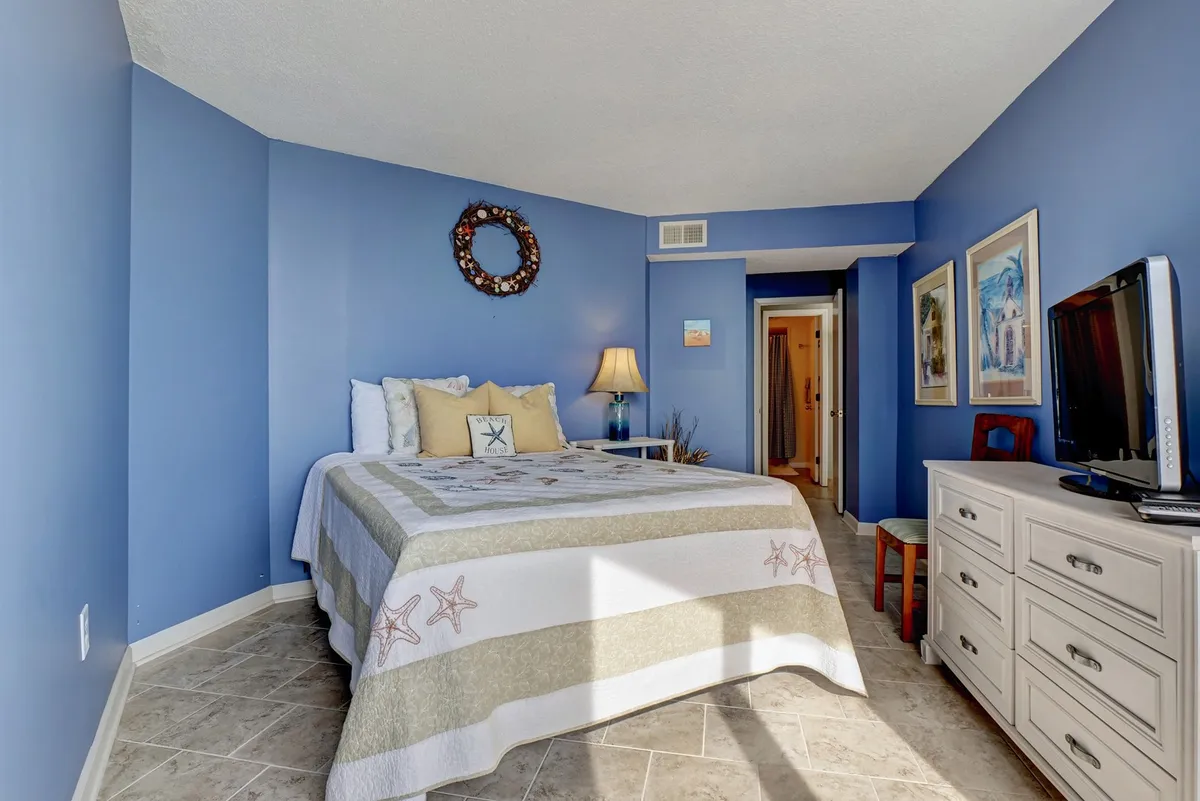 A tranquil bedroom featuring a bed with decorative bedding, calming wall art, a warm lamp, and a classic dresser. Soft natural light filters through the window, creating a peaceful and private atmosphere ideal for relaxing and unwinding.