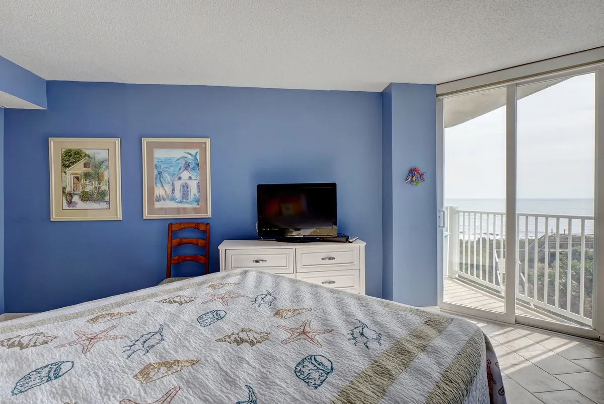A cozy bedroom with a queen-sized bed dressed in a patterned quilt, a television, wall art, and a sliding glass door that opens to direct ocean views. The room is bathed in natural light and offers a tranquil setting for rest and relaxation.