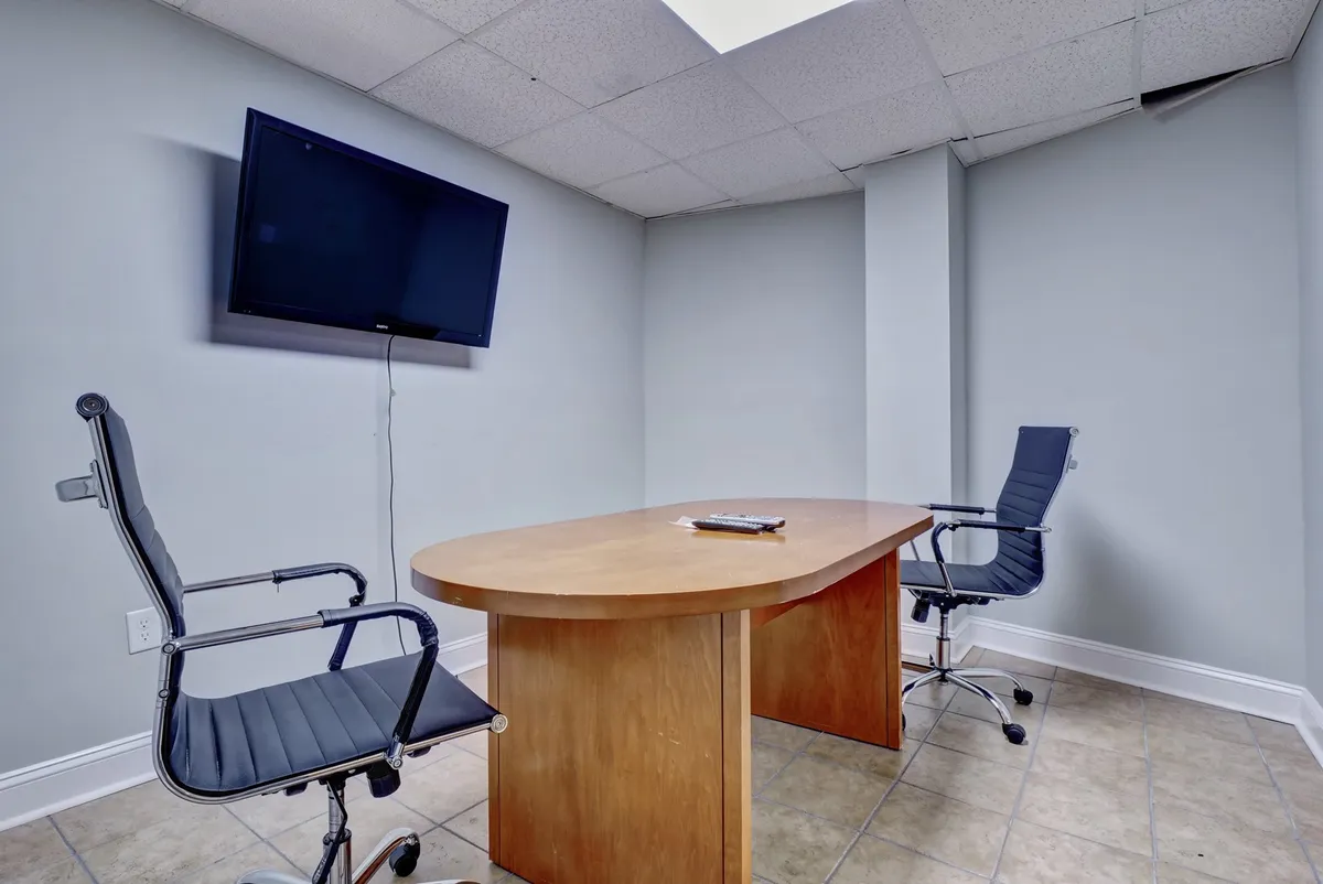 A functional business center interior, featuring a large wooden conference table, two ergonomic office chairs, and a flat-screen TV. The setup suggests a professional space suitable for meetings, video calls, or focused work sessions, all within the resort.