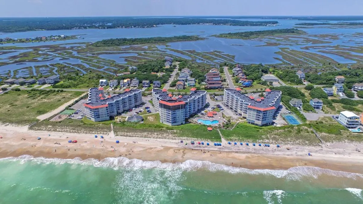 A wide-angle exterior shot showing beachfront resort buildings, a large swimming pool with blue umbrellas, and the sandy shoreline with ocean waves just beyond. Families can be seen enjoying the pool, while others are gathered near the beach, highlighting the close connection between resort amenities and the natural oceanfront setting.