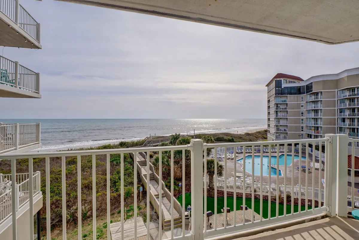 A serene exterior view looking over a sparkling pool with the ocean and sandy beach beyond. The balcony railing in the foreground gives a sense of vantage, suggesting the viewer is standing on a raised deck with panoramic views of the pool and the sea horizon.