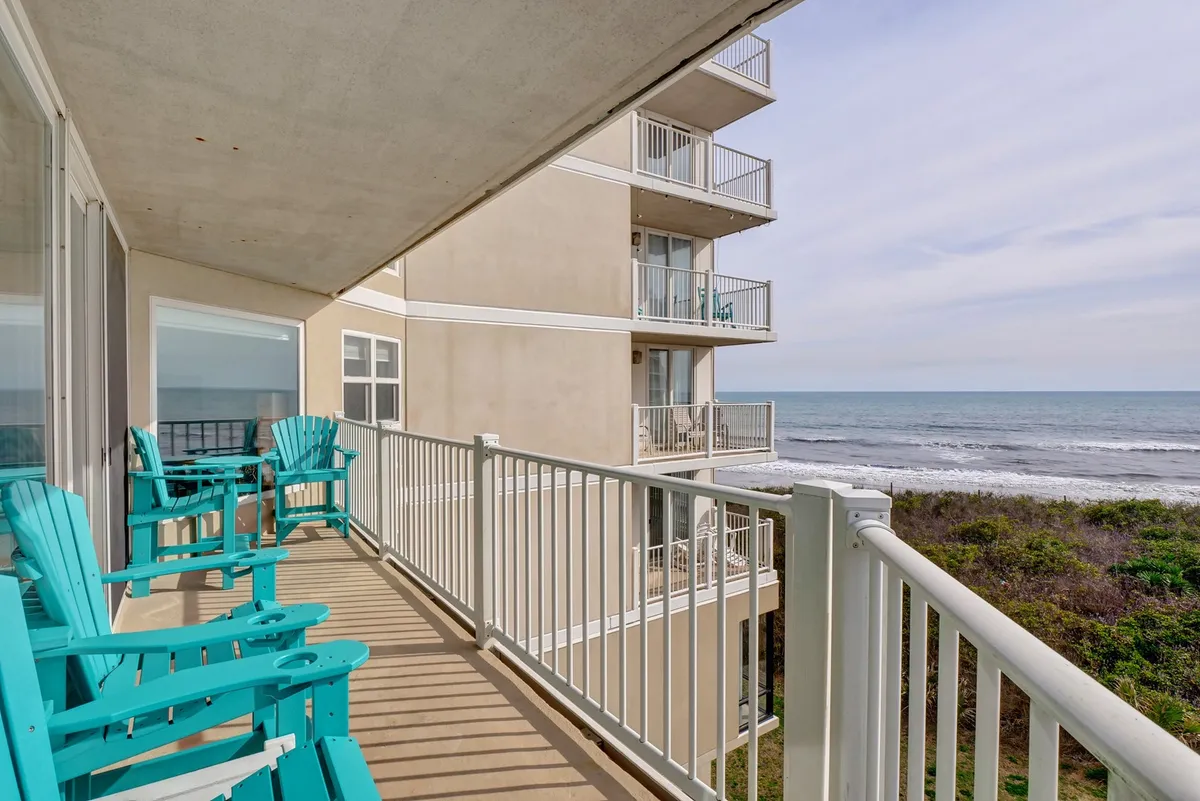 This image shows a tranquil balcony or deck setup, featuring turquoise Adirondack chairs facing an expansive ocean view. The balcony railing frames the scene, suggesting a private, covered deck space perfect for relaxation. The lighting indicates either morning or evening, enhancing the atmosphere of serenity and escape.