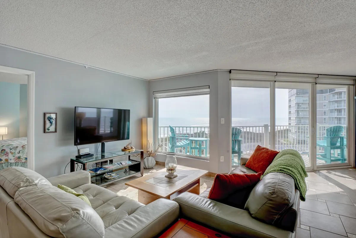 A spacious living area with comfortable seating arranged to face large windows that offer sweeping ocean views. The setup includes a modern TV, a decorative coffee table, and plenty of natural light filling the room, creating an inviting space for relaxation or group activities.