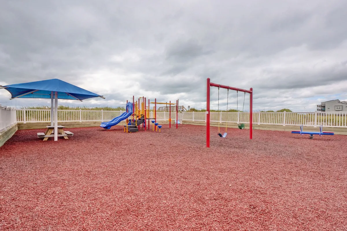 A vibrant playground area with colorful equipment and swings, surrounded by a picnic area where families gather. Children are happily playing, and the atmosphere is filled with laughter and energy. The setting is open and bright, offering a safe, joyful space just steps from the accommodations.